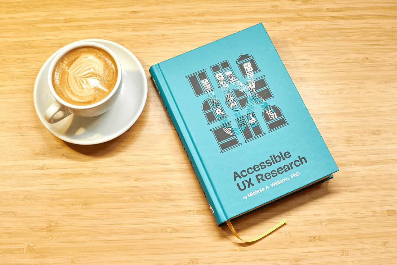 Hardcover edition of Michele Williams’ new book ‘Accessible UX Research’ on a wooden table, with a cup of coffee next to it. The book has a teal cover that shows a three times three grid of windows in different architectural styles. On top of the windows, there’s a light varnish that spells out the word ‘UX’ with research-related icons such as speech bubbles, a looking glass, a keyboard, and UI components.