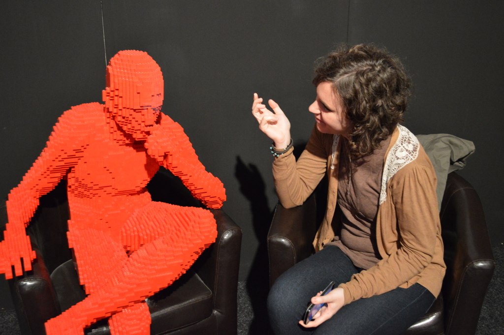 A real person sits in a chair pretending to interview another person made out of orange legos sitting in a chair next to them.